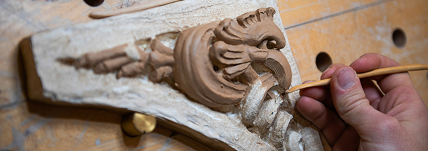 A closeup of a hand shaping a carving of a cockatrice using a clay-sculpting tool.