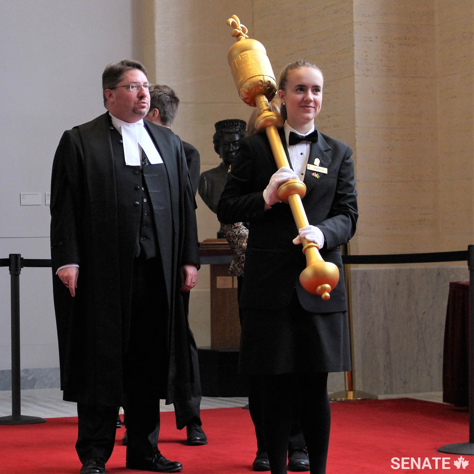 Senate Page Amélie Boutin prepares to carry the mace into the Senate Chamber as model Speaker Adam Thompson — normally a Senate table officer — looks on. The Fédération de la jeunesse canadienne-française generously loaned out its mace for the occasion. The real Senate cannot meet without its mace being present.