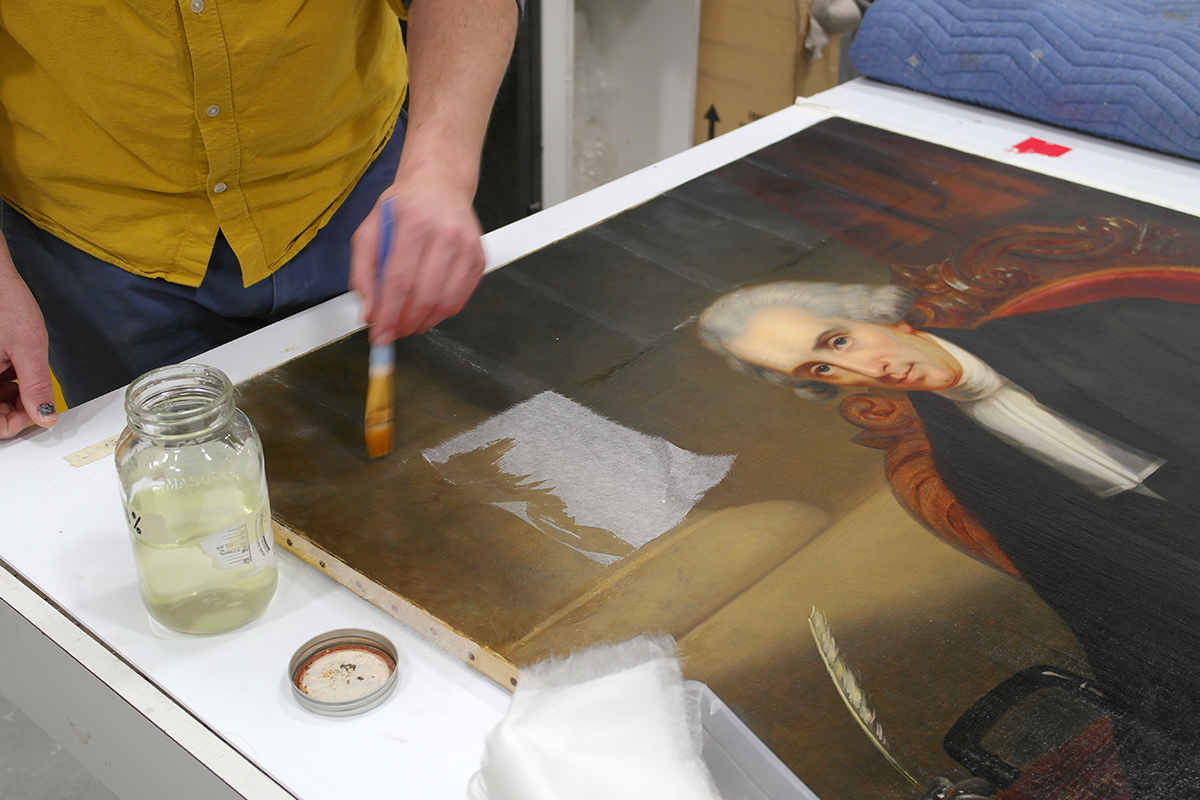A conservator applies facing tissue to protect the painting before beginning work on the back of the canvas. (Photo credit: Legris Conservation)