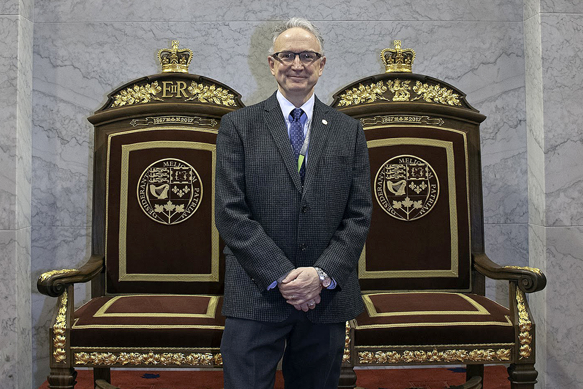 M. White pose avec les trônes royaux qu’il a conçus pour la Chambre temporaire du Sénat, qui se trouvent dans l’édifice du Sénat du Canada. Les détails de style beaux-arts qui ornent les trônes sont inspirés de l’édifice, bâti en 1912, qui abritait à l’origine une gare ferroviaire. (Crédit photo : Services publics et Approvisionnement Canada)