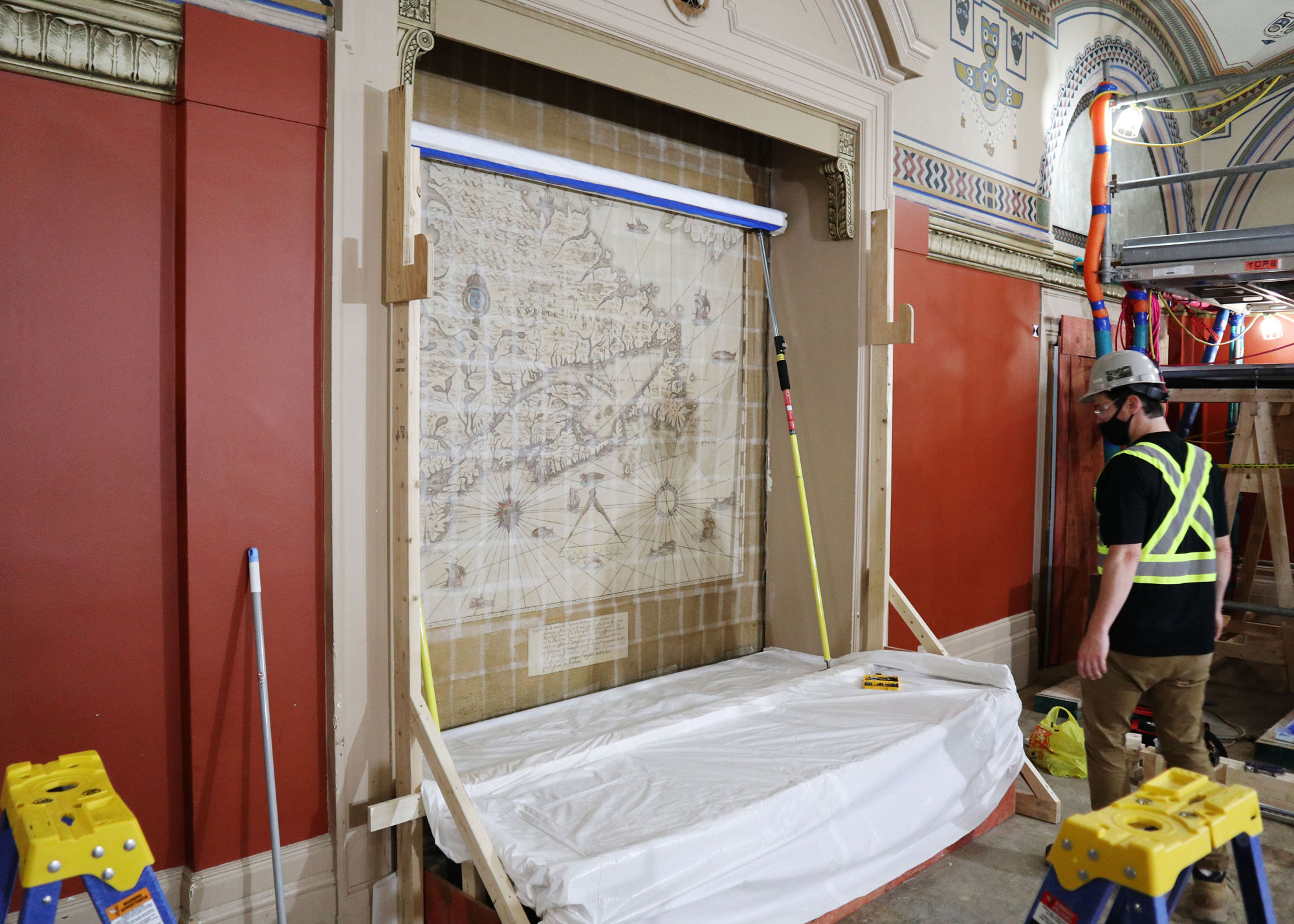 Des travailleurs préparent une murale représentant une carte, dans la Salle de comité des chemins de fer du Sénat, afin de la retirer du mur. On peut voir, à droite, les échafaudages qui ont été installés pour retirer les murales qui étaient collées en haut des murs. (Crédit photo : Legris Conservation Inc.)