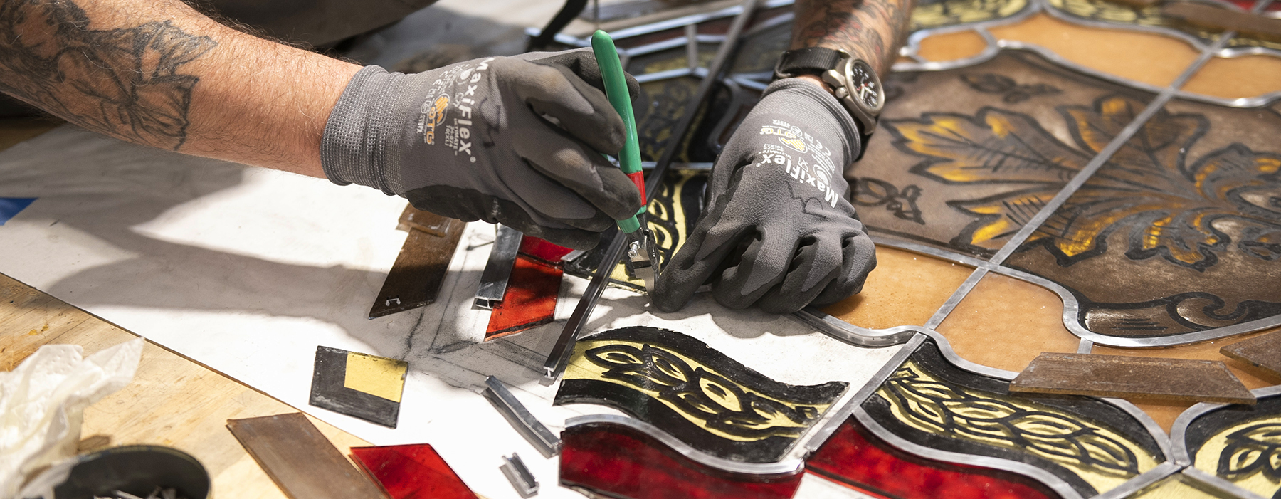 A conservator with gloved hands uses a tool to replace strips of lead that hold panes of stained glass in place.
