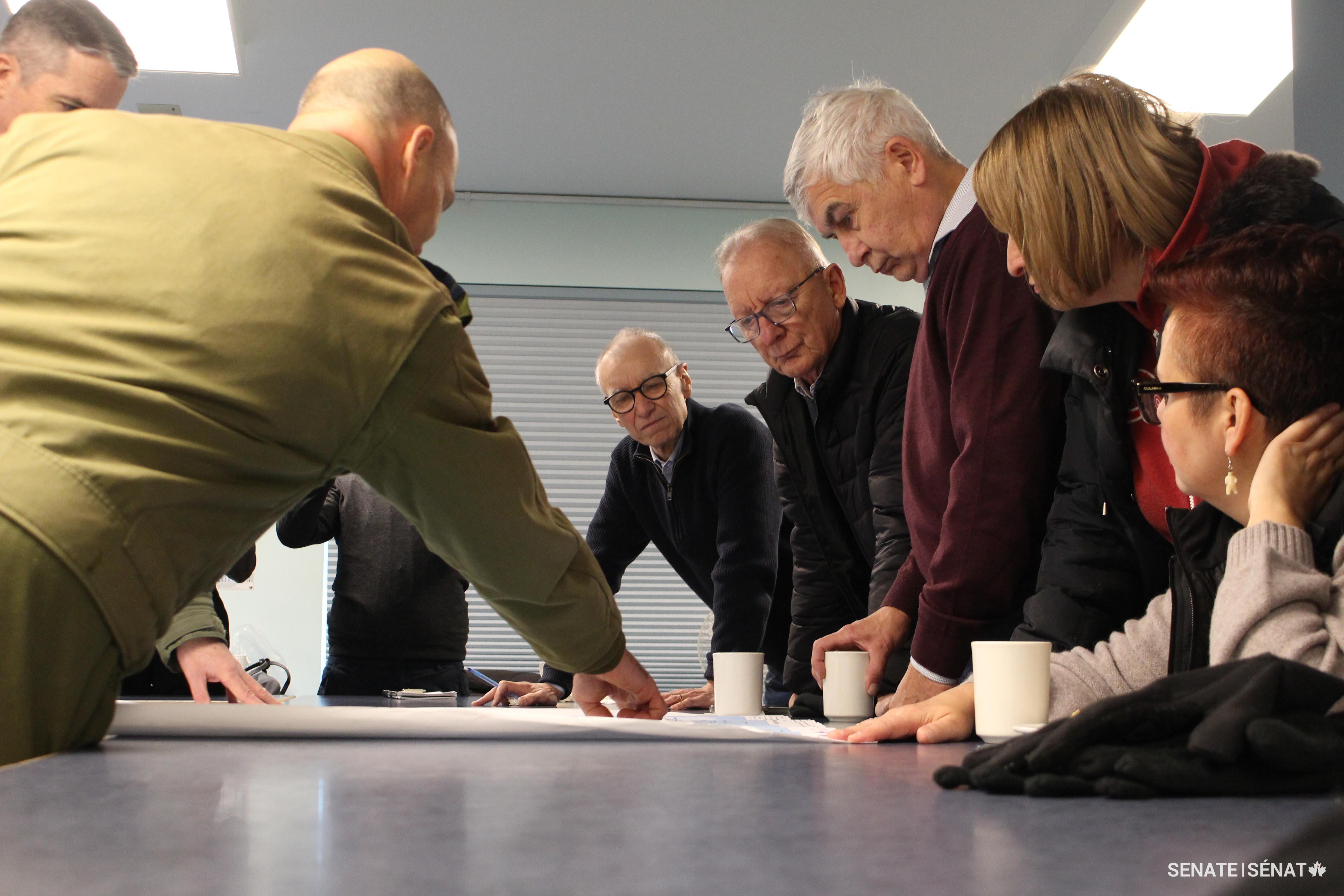 De droite à gauche, les sénateurs Anderson, Dasko, Gignac, Boisvenu et Dean examinent une carte de l’Arctique avec des membres des Forces armées canadiennes dans un réfectoire à Inuvik, dans les Territoires du Nord-Ouest.