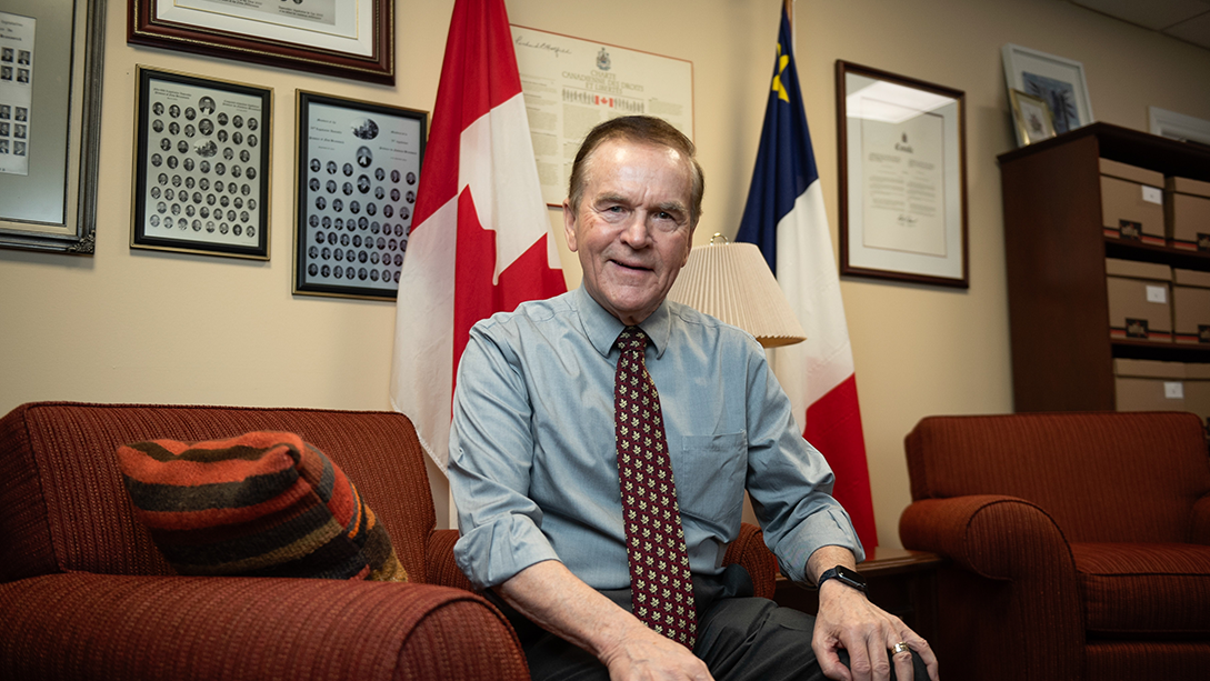Le sénateur Percy Mockler est assis dans un fauteuil, dans son bureau, devant les drapeaux du Canada et de l’Acadie.
