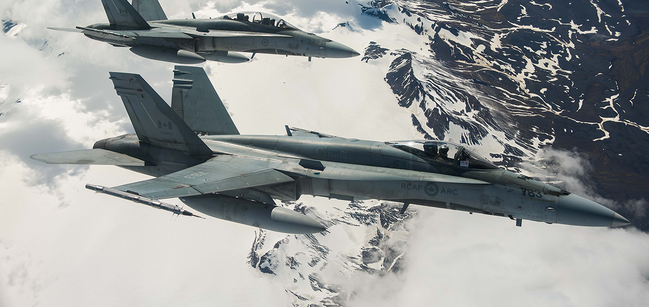 Two grey fighter jets from the Royal Canadian Air Force fly over a snowy mountain range.