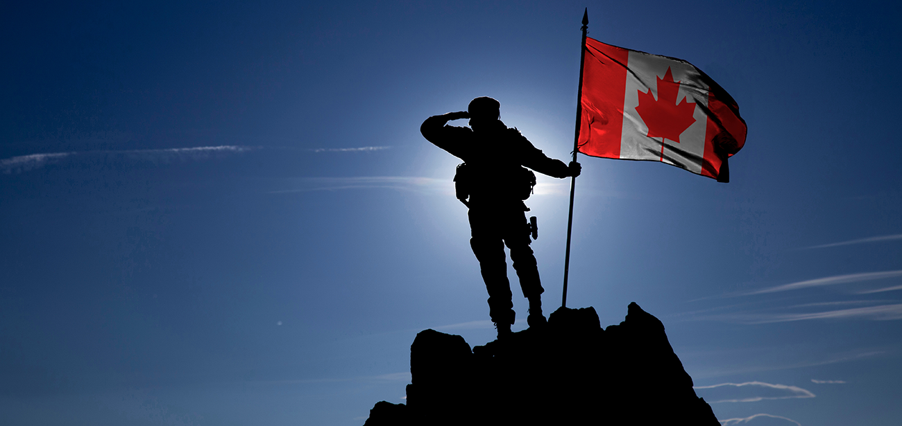 The backlit silhouette of a soldier standing on the top of a mountain, holding a Canadian flag hoisted on a flagpole.