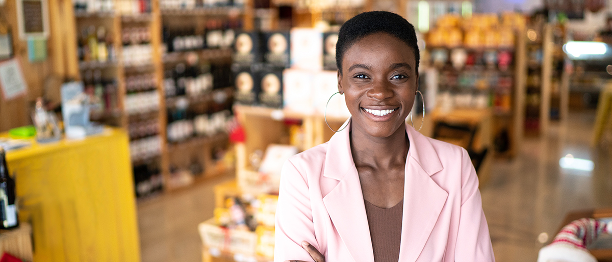 Une femme noire souriante et portant un tailleur se tient au premier plan d’un magasin.