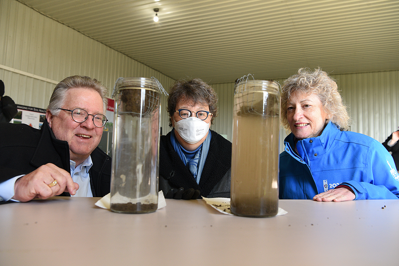 Les sénateurs Rob Black, Paula Simons et Pat Duncan observent les résultats d’un test de stabilité du sol dans deux bocaux de verre.